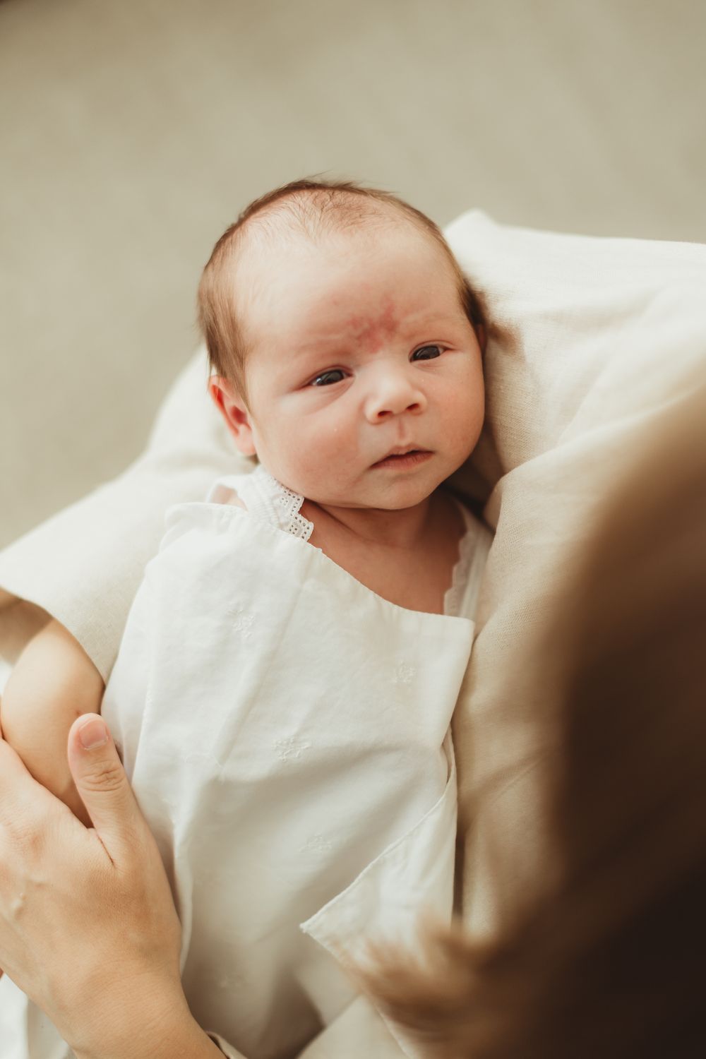 A newborn baby wrapped in a white blanket being held by a parent, looking alert with dark hair.