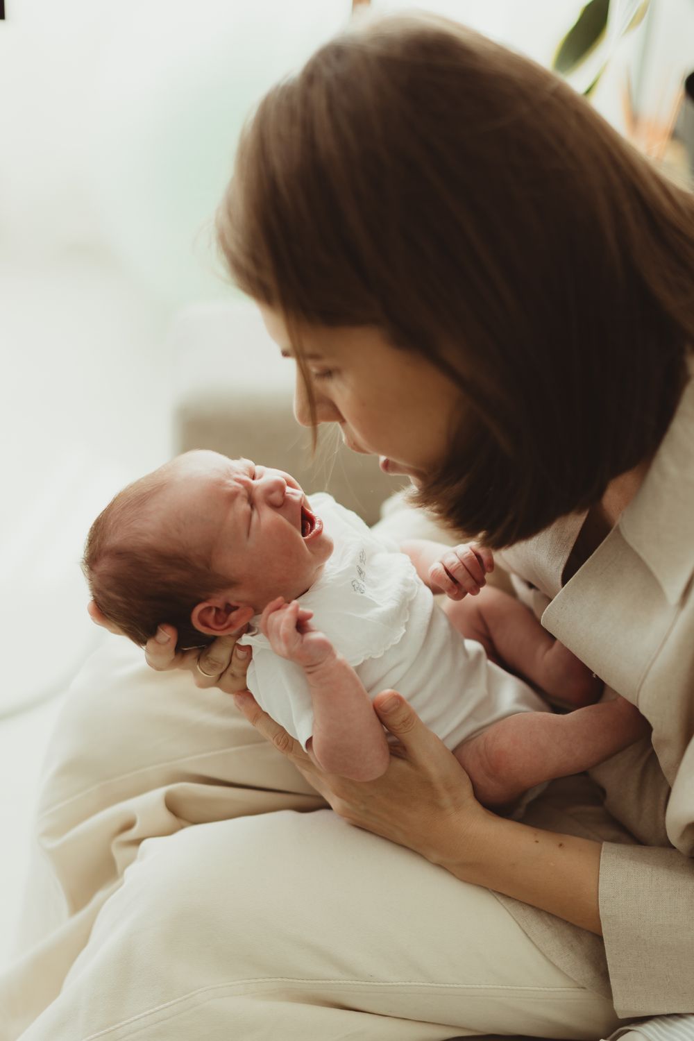 A mother tenderly holds her crying newborn baby in white clothing while sitting on a light-colored sofa.