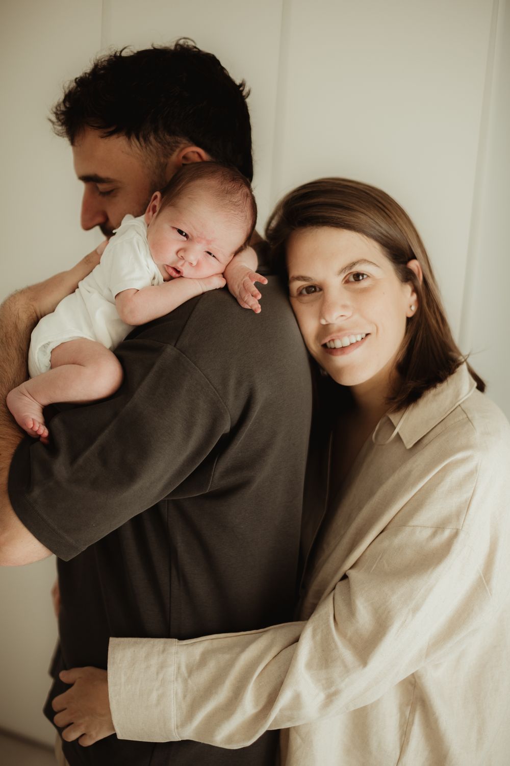 A smiling mother and father hold their newborn baby together in a warm, intimate family portrait.