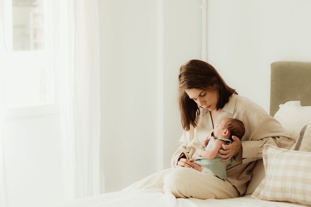 A young mother sits on a bed cradling her newborn baby in a bright, minimalist white bedroom.