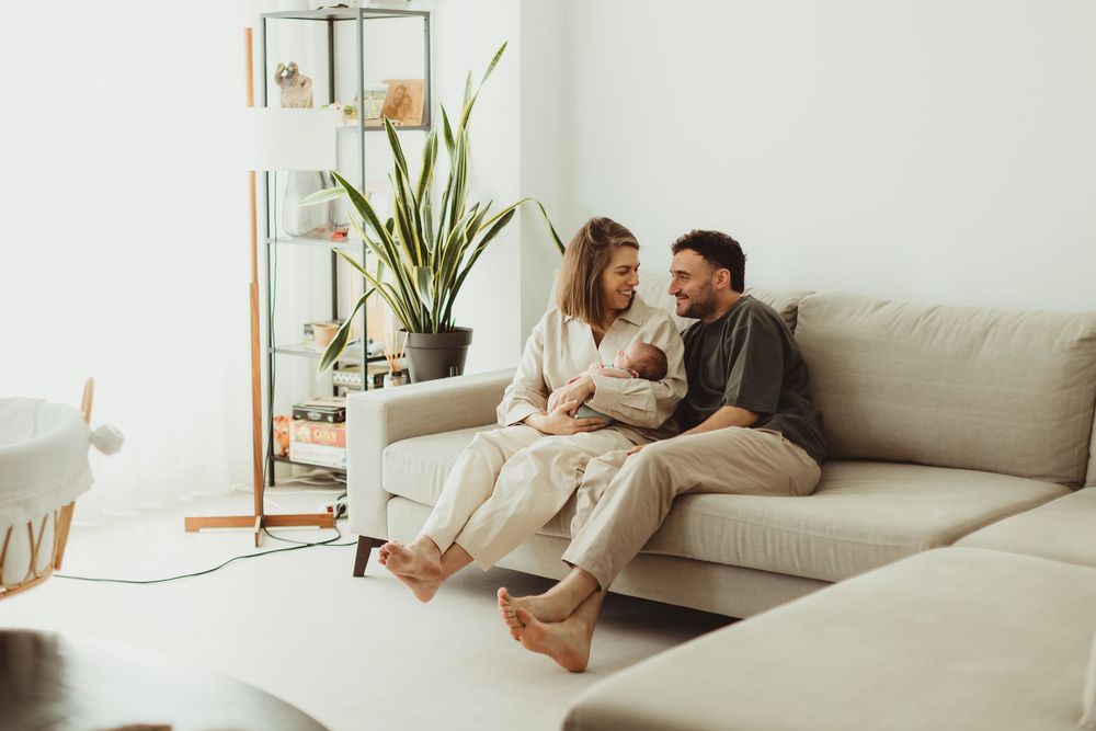 Happy couple sitting on a beige sofa holding their newborn baby in a bright modern living room.