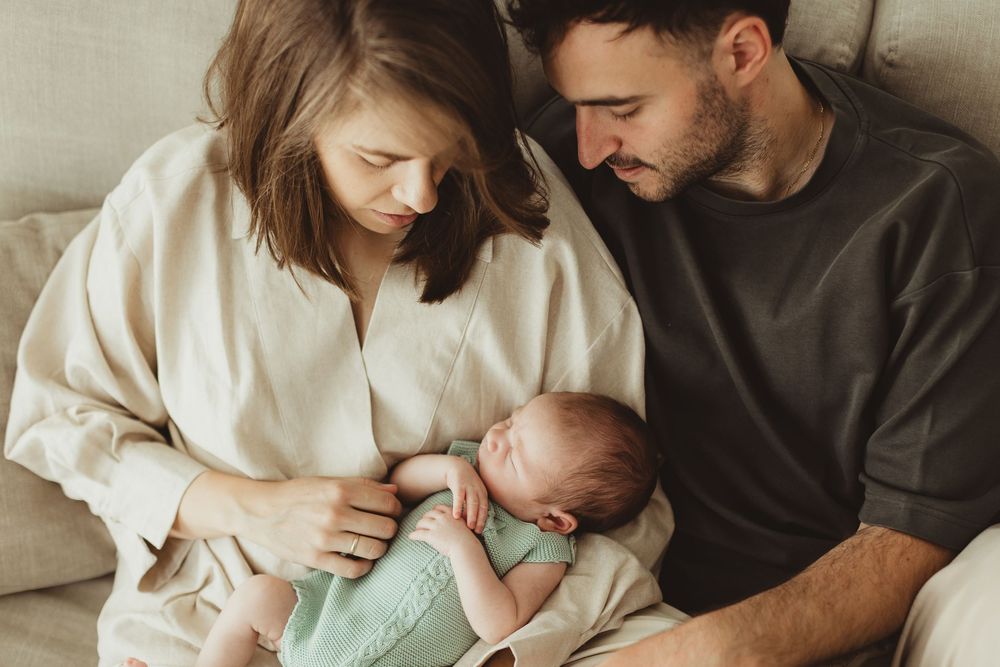 New parents sitting on a couch together, lovingly gazing at their sleeping newborn baby wrapped in a mint green blanket.