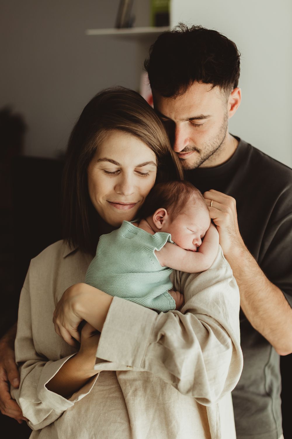 New parents tenderly holding their sleeping newborn baby wrapped in a mint green blanket at home.