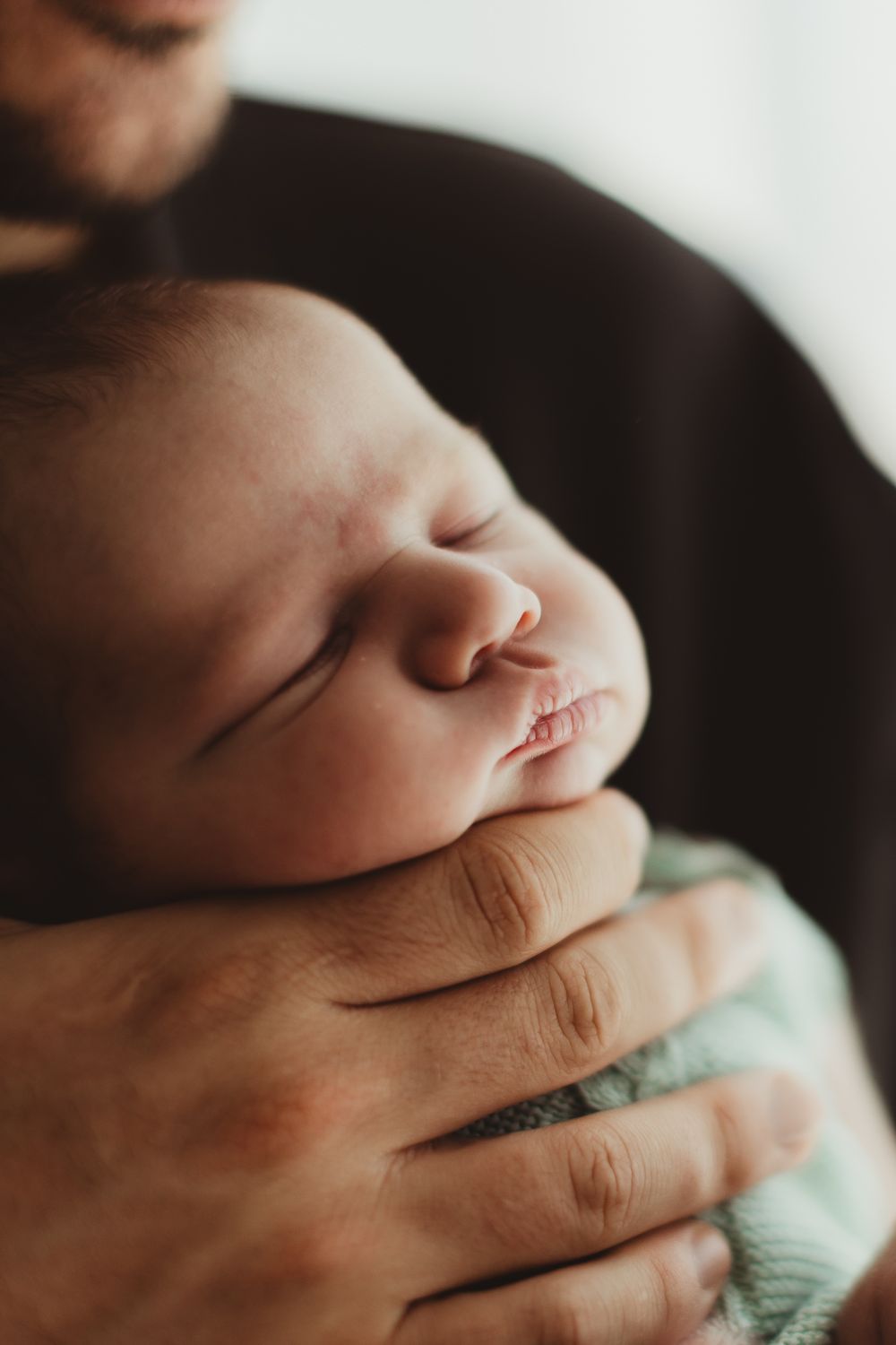 Newborn baby sleeping peacefully in father's arms, wrapped in a soft mint green blanket.