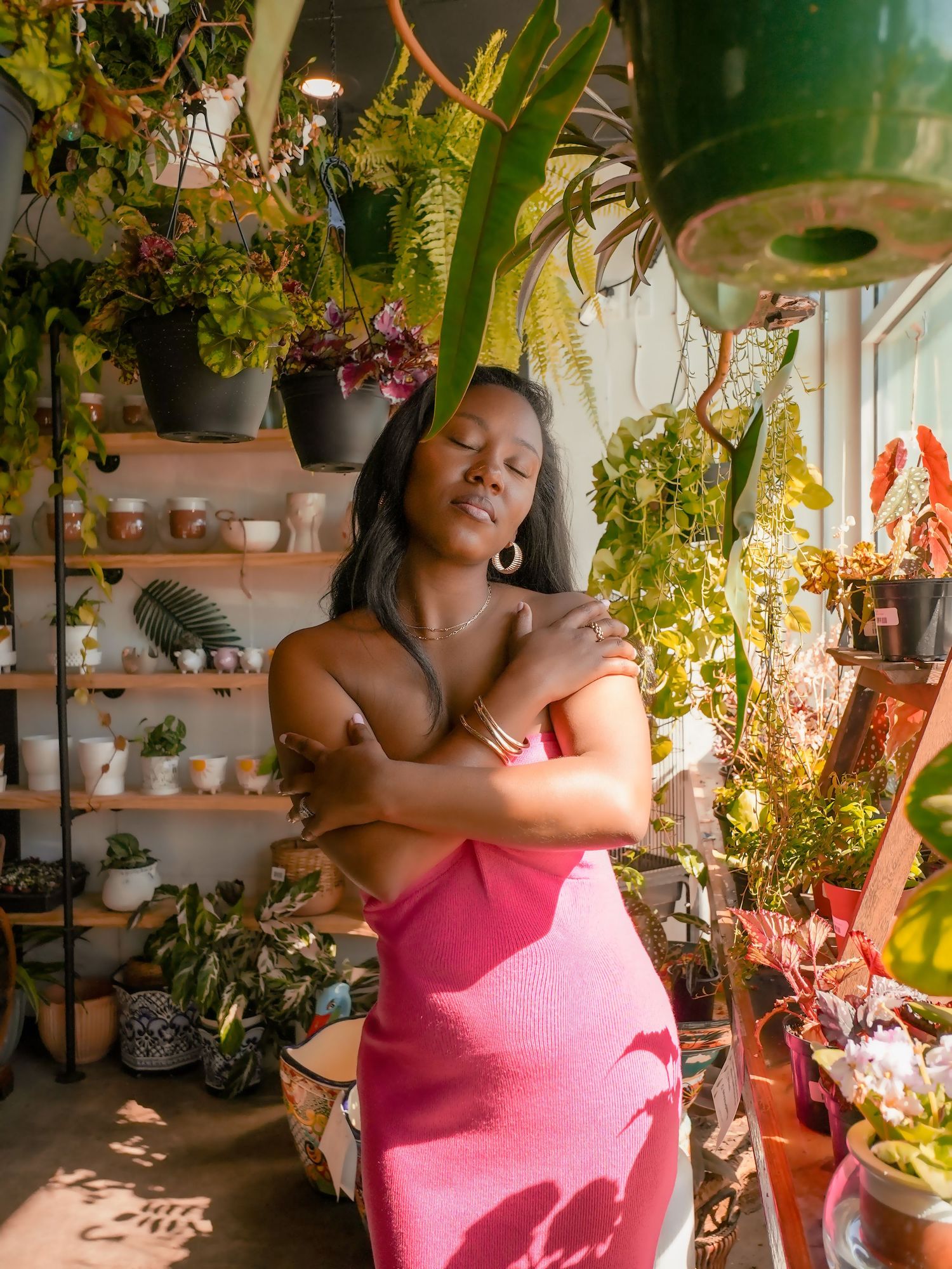 Figure in pink dress stands surrounded by potted plants in a sunny indoor garden space.