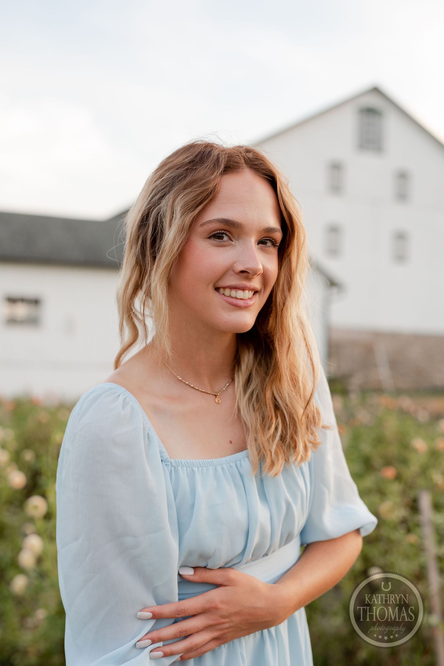 A soft blue dress creates a romantic look against a white barn backdrop with blooming flowers.