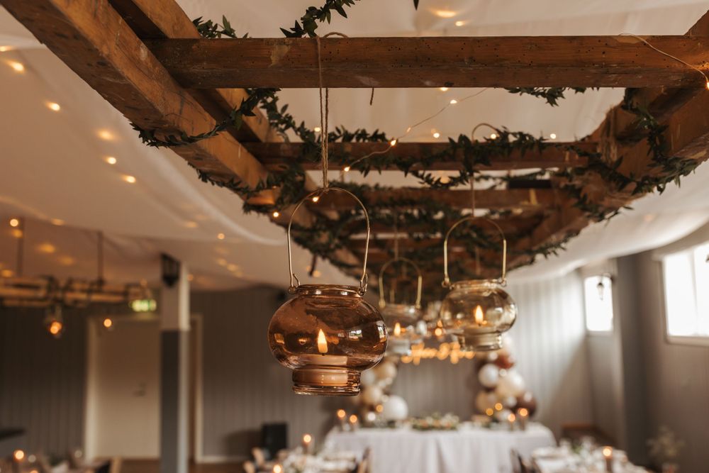 Modern reception venue with wooden beam ceiling decorated with hanging metallic orbs and greenery.