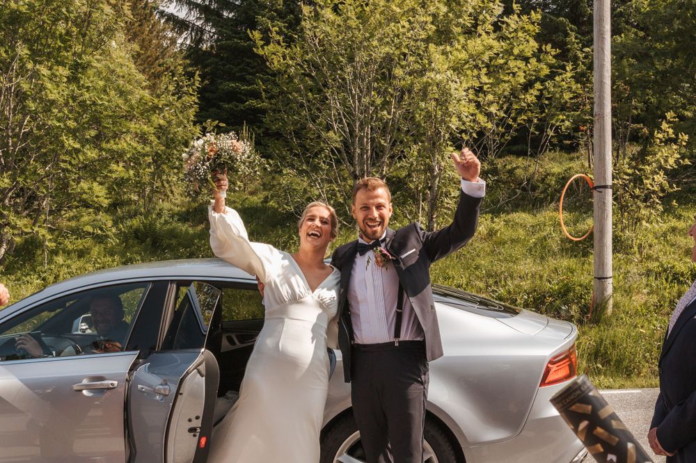 Newlyweds celebrate with raised arms next to silver car outdoors.