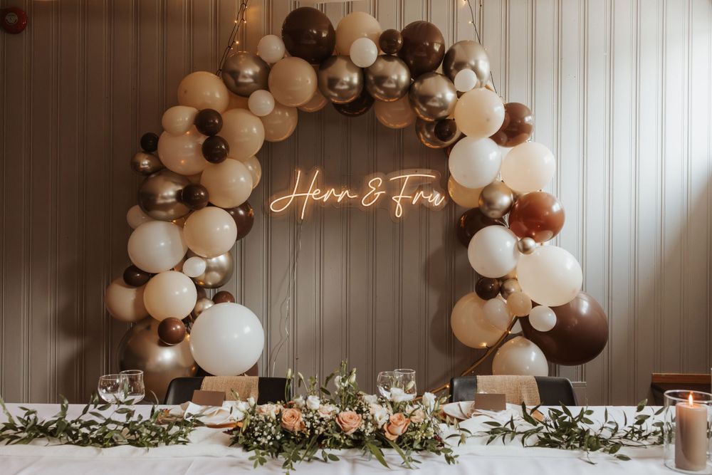 Brown and white balloon arch with neon sign centerpiece and floral decorations.
