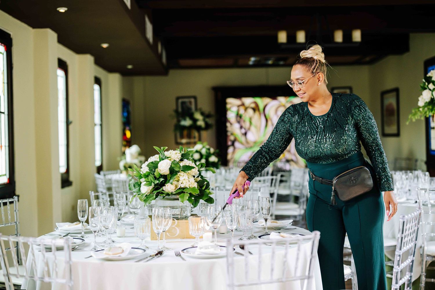 Event staff member in dark green dress arranging place settings at wedding reception.