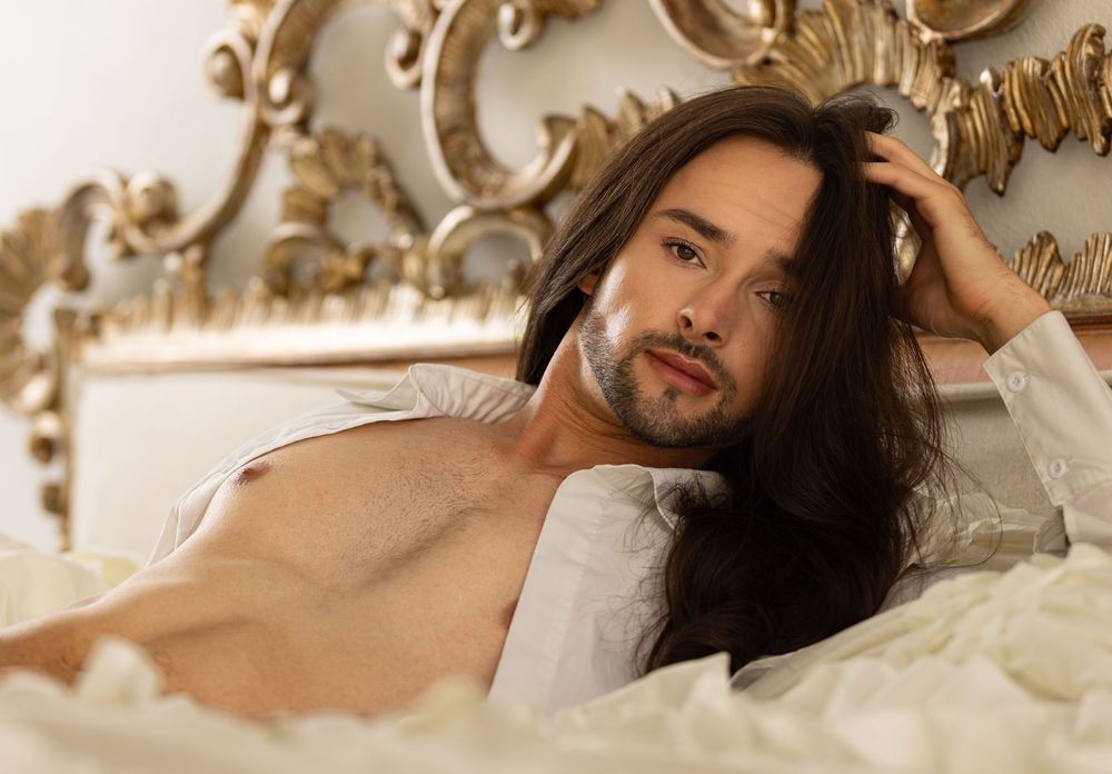 Man with long hair and open shirt lying in bed during boudoir photo shoot in Los Angeles photography studio.