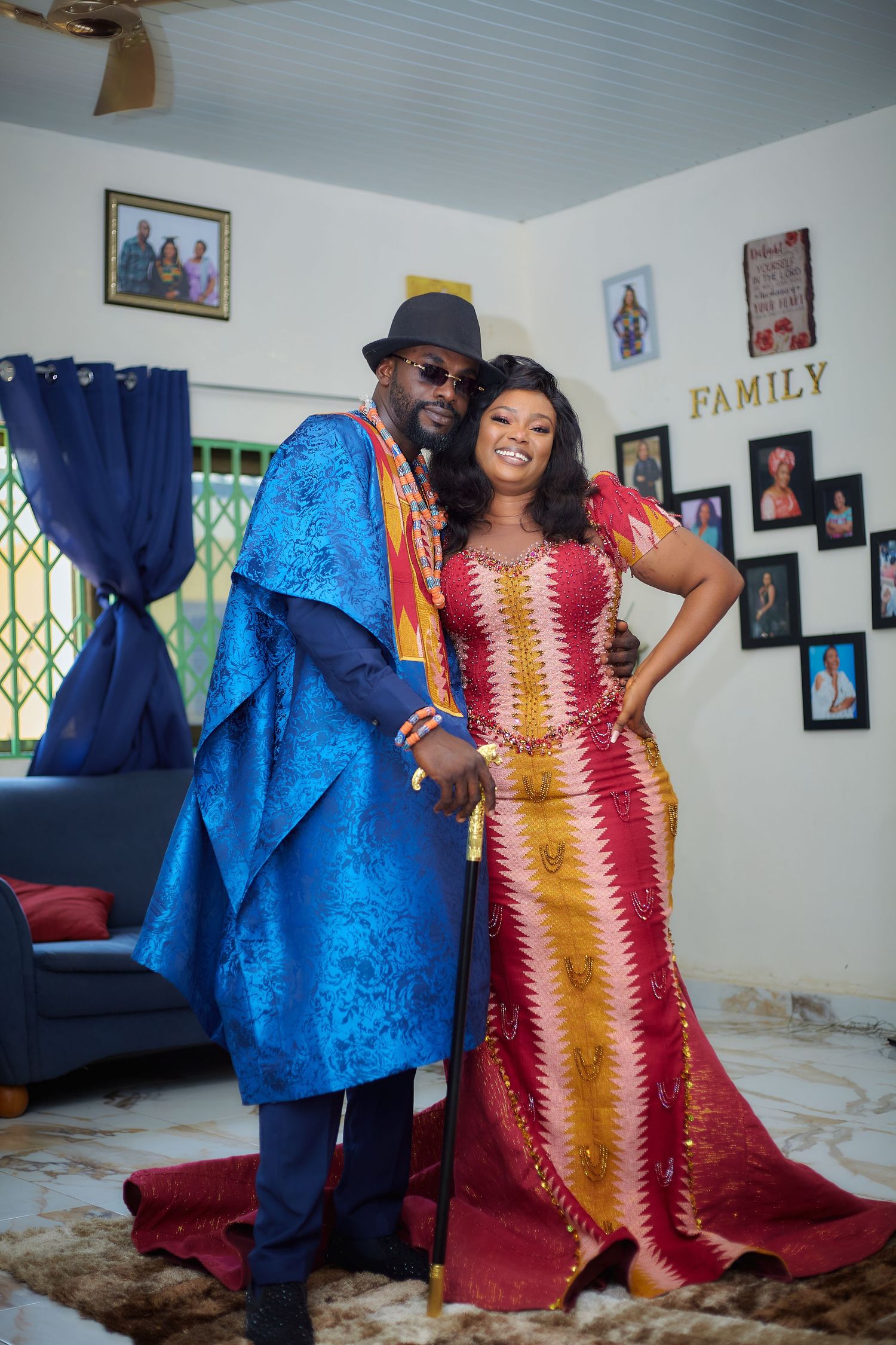 Couple in blue agbada and red kente dress dancing joyfully indoors.
