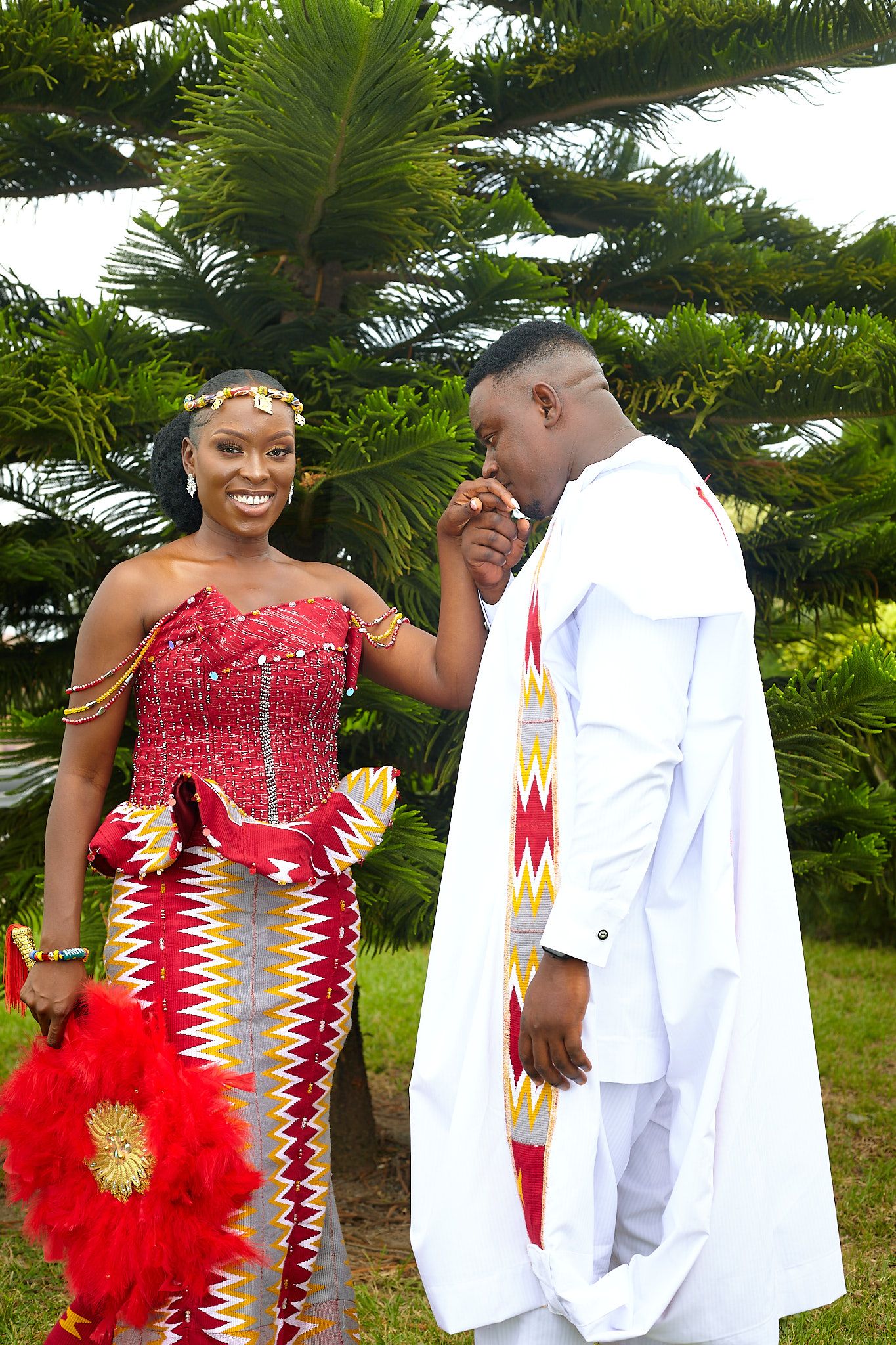 Couple in traditional African attire posing outdoors with red kente dress and white agbada.