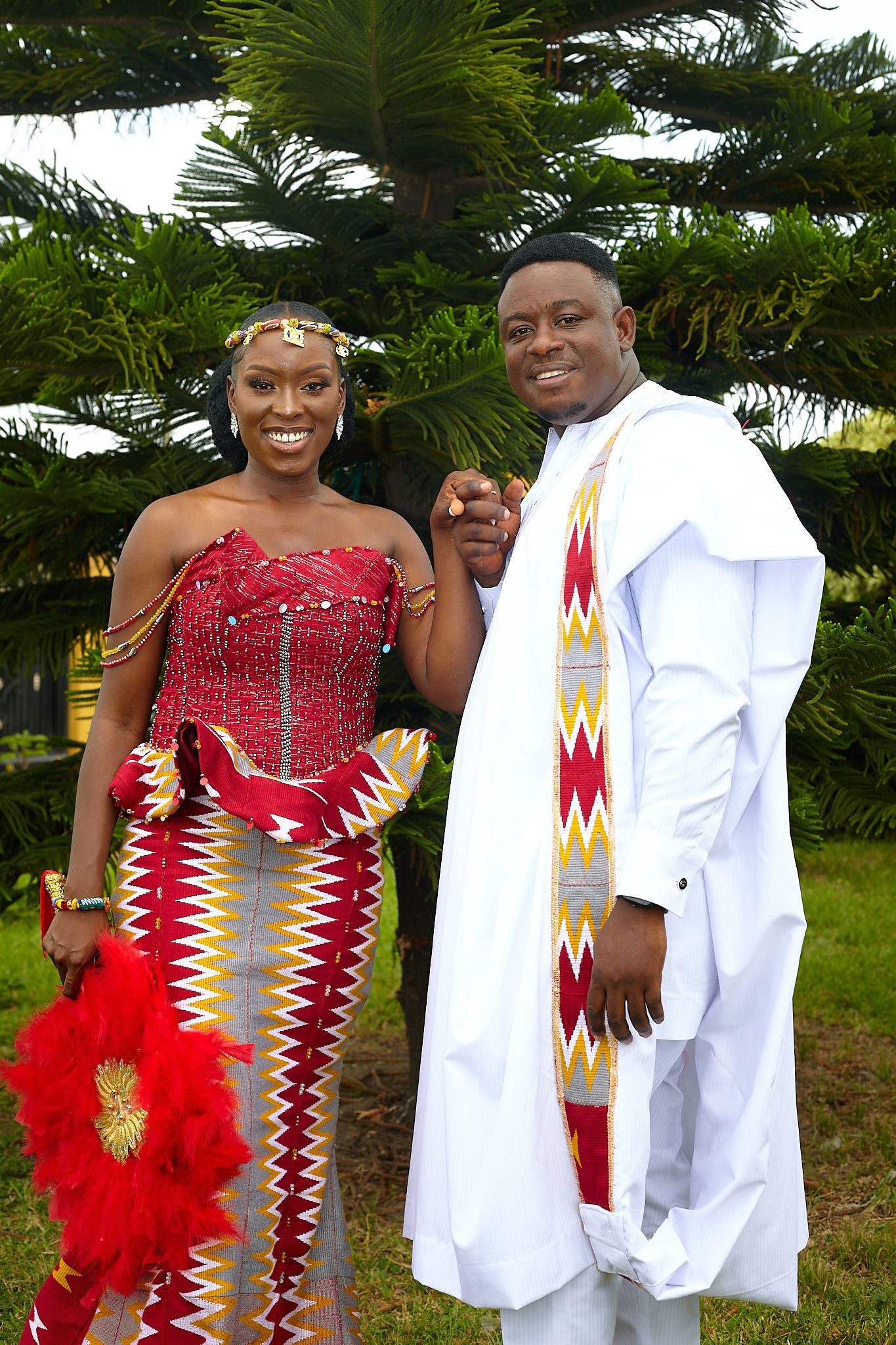 Wedding couple in red kente dress and white agbada smiling together against palm trees.