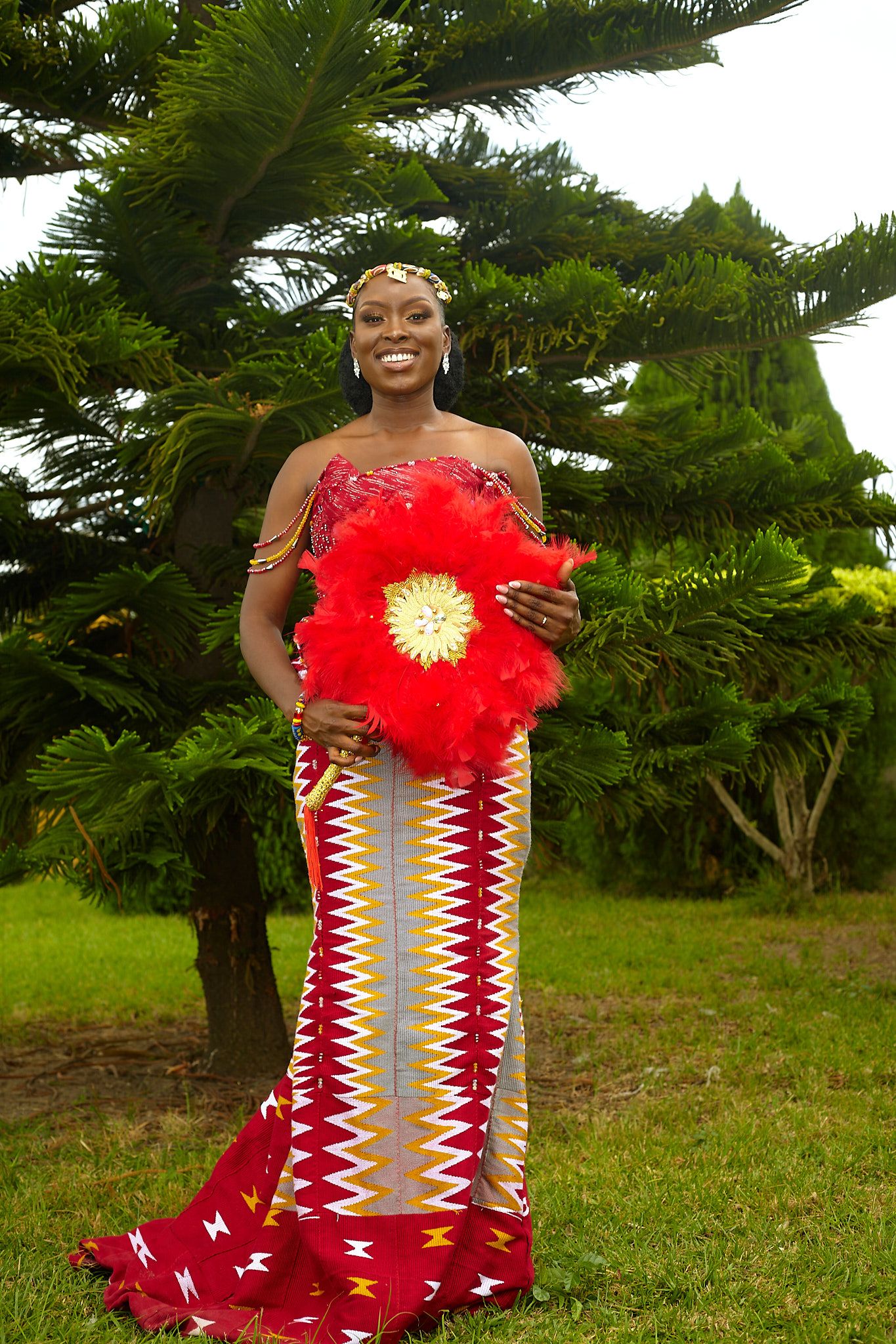 Woman in red traditional dress with feather detail poses in outdoor garden setting.