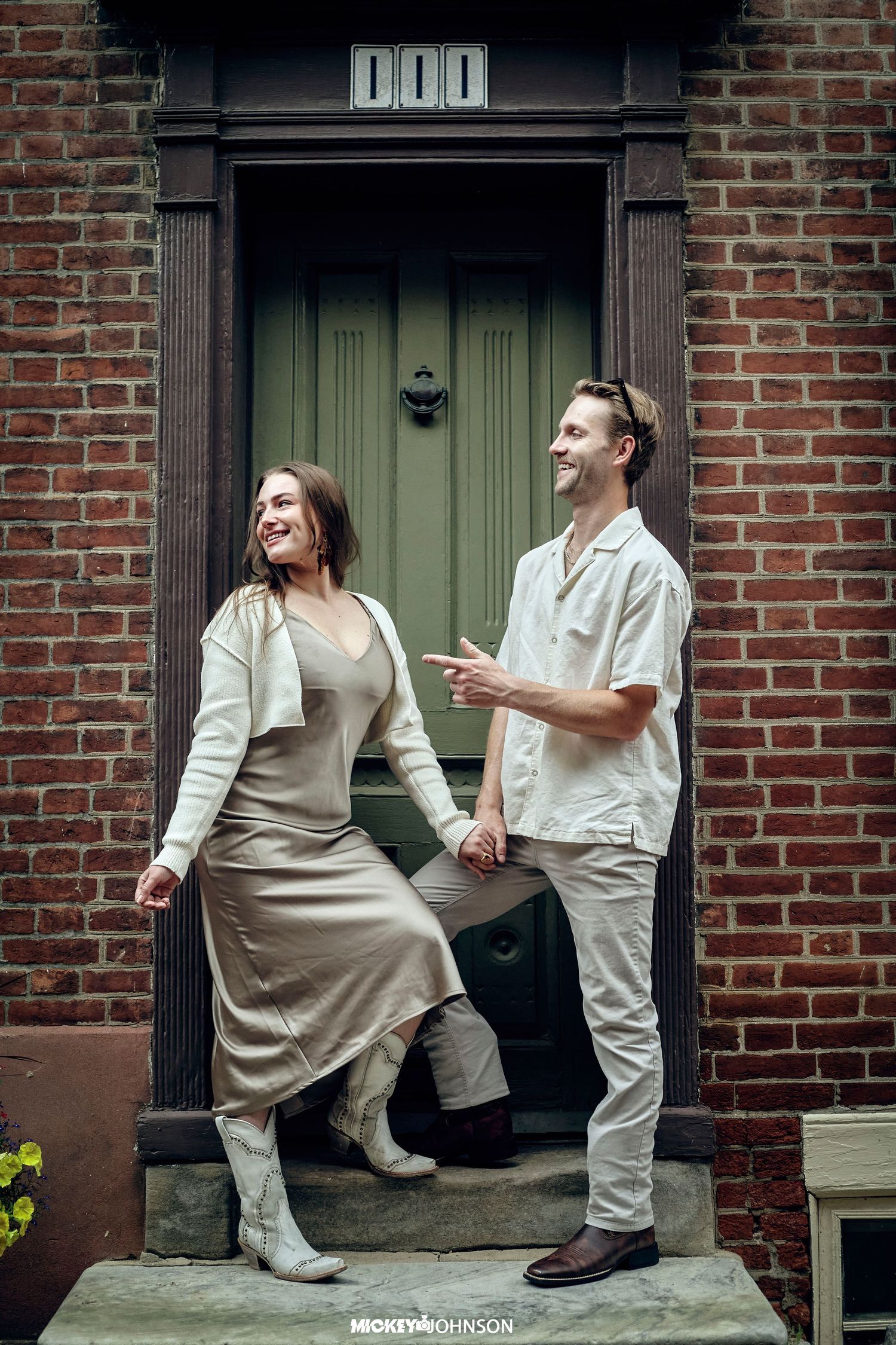 Two people in light-colored outfits pose playfully against a brick wall exterior.