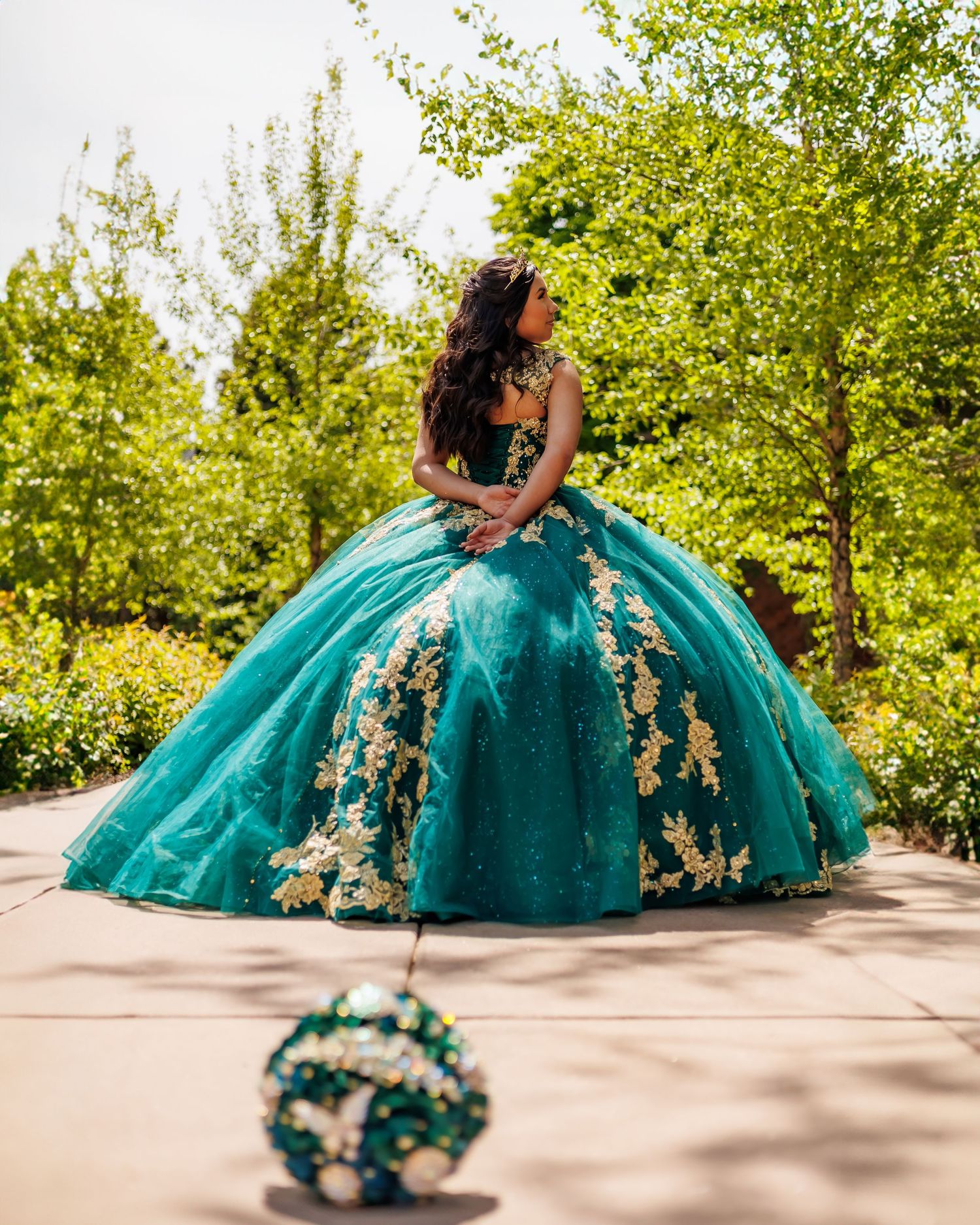 Stunning turquoise quinceañera ball gown with floral appliques photographed in garden setting.