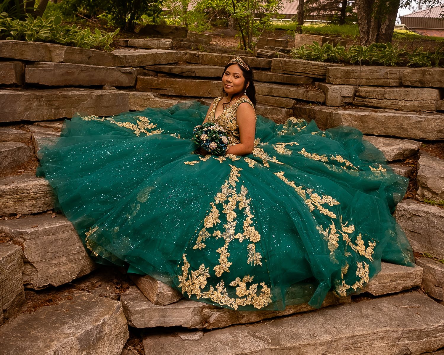 A stunning teal quinceañera gown with gold embellishments photographed on stone steps in an outdoor setting.