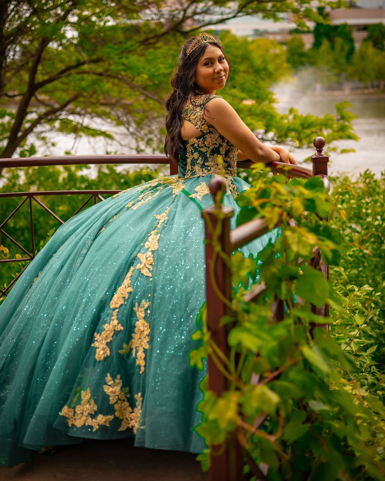 Elegant teal ball gown with gold appliques sparkles in a garden setting against green foliage.