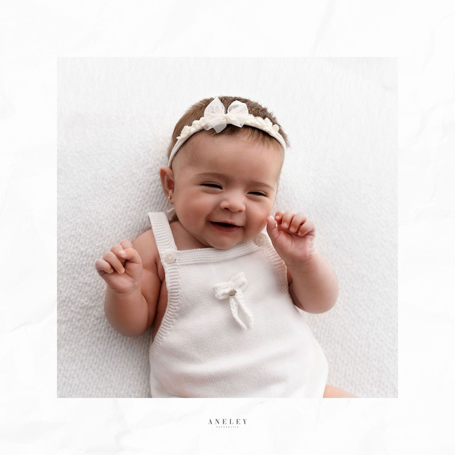 Joyful baby in white outfit with bow headband smiles during portrait session.