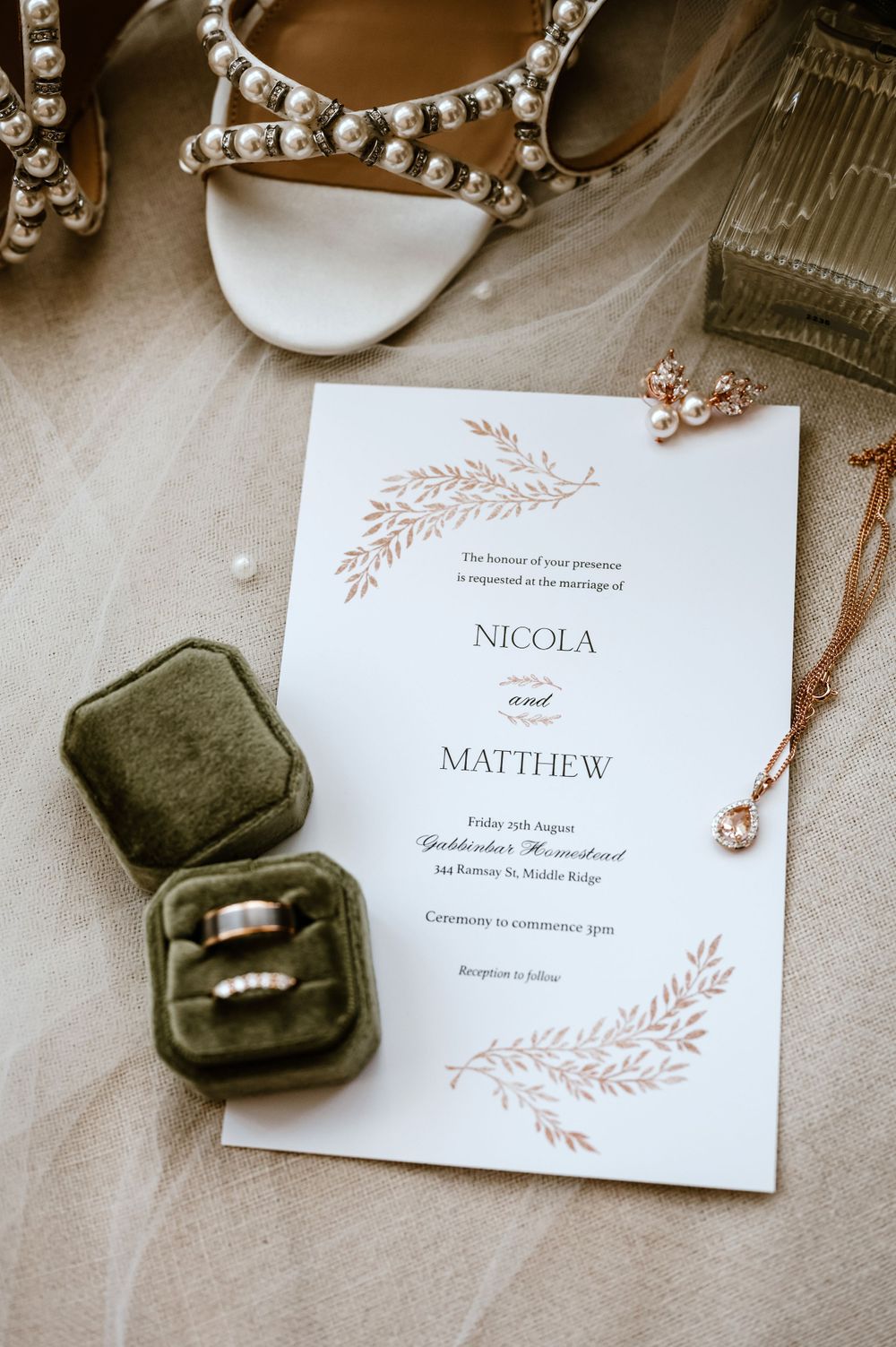 Wedding invitation with floral design alongside pearl shoes, rings in green velvet box, and jewelry.