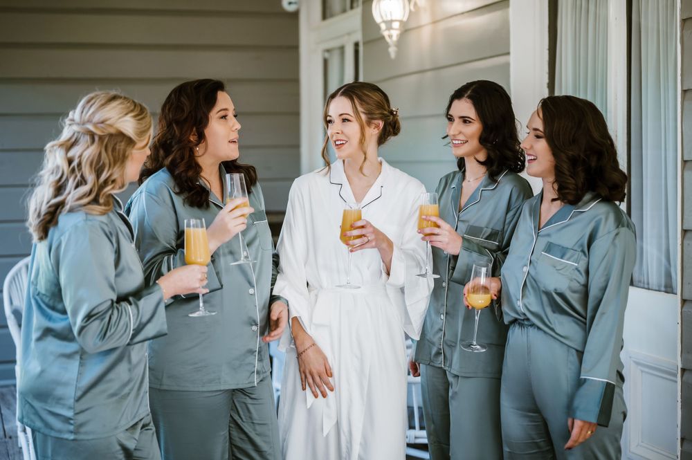 A group in matching blue silk pajamas shares drinks and laughter during wedding preparations.