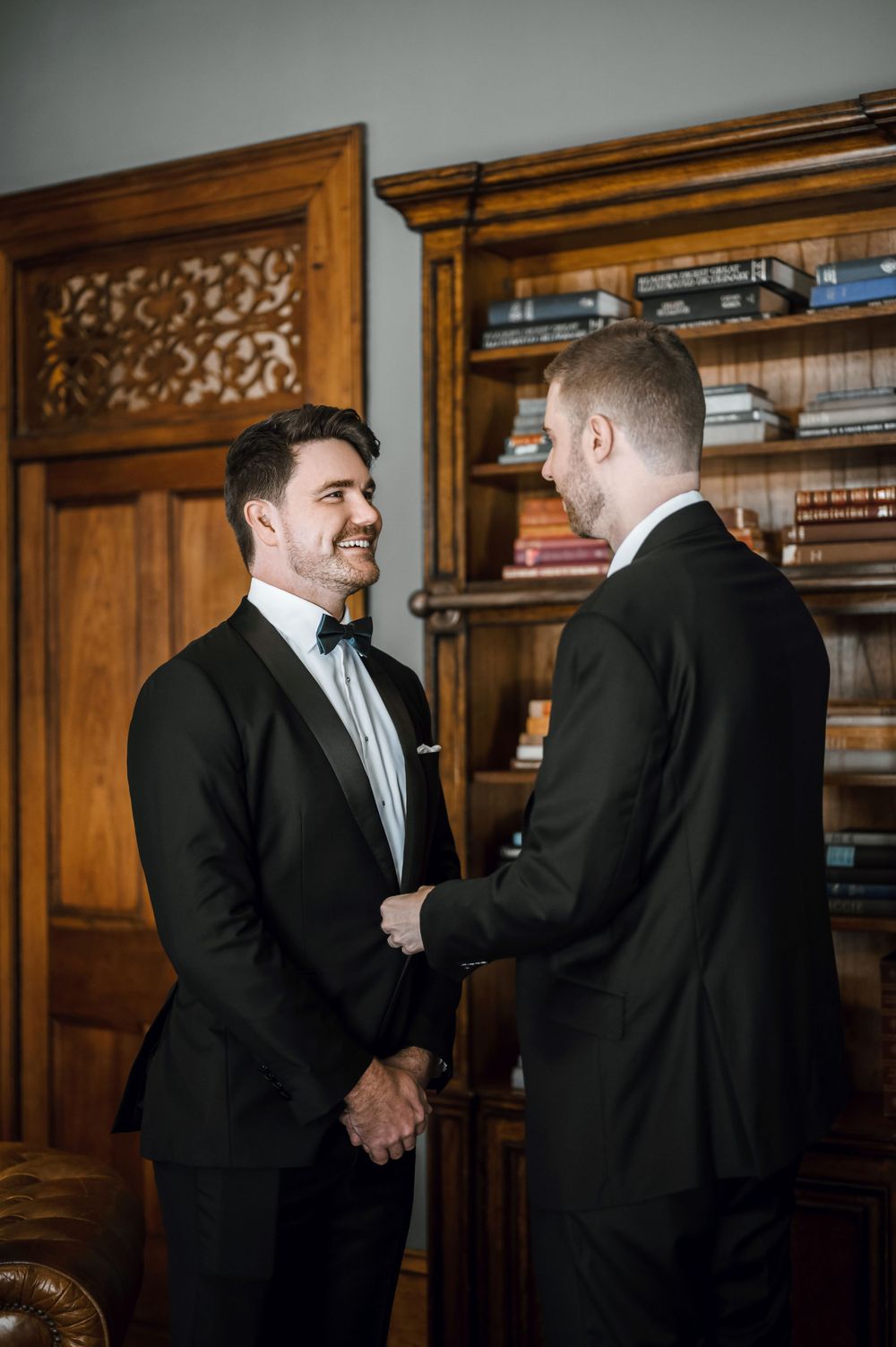 Two formal-dressed men share a candid moment of laughter in front of wooden bookshelves.