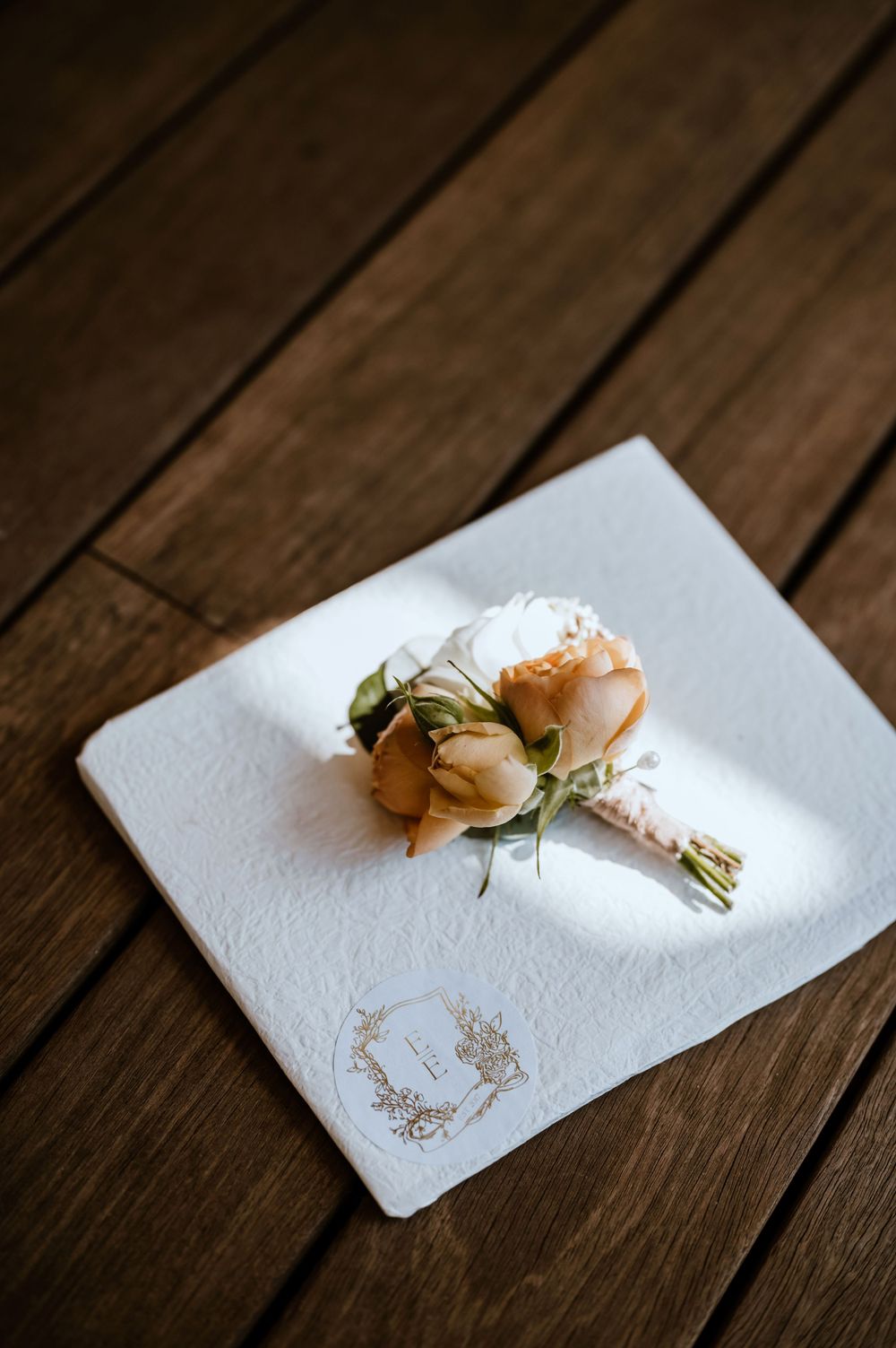 Small delicate peach-colored boutonniere rests on white textured napkin against dark wooden surface.