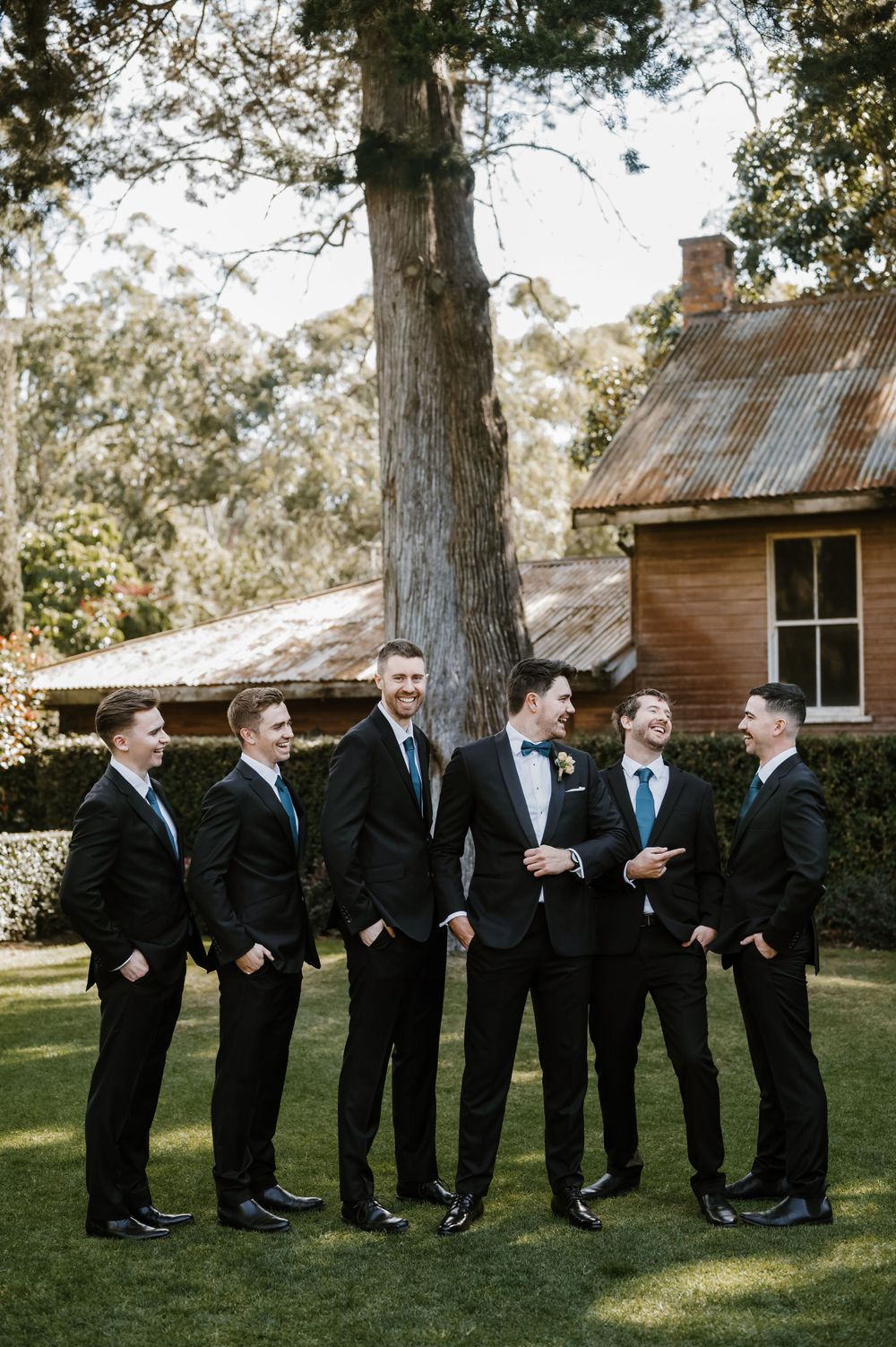 Wedding party in black suits gather on a lawn in front of a rustic building.