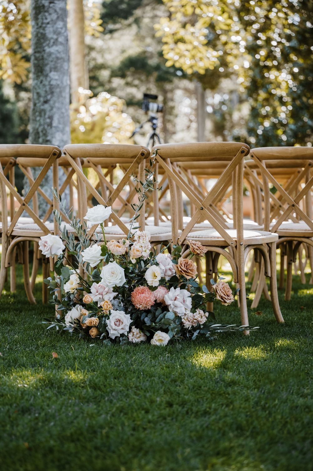 Elegant floral arrangement adorns outdoor wedding ceremony seating with cross-back chairs.