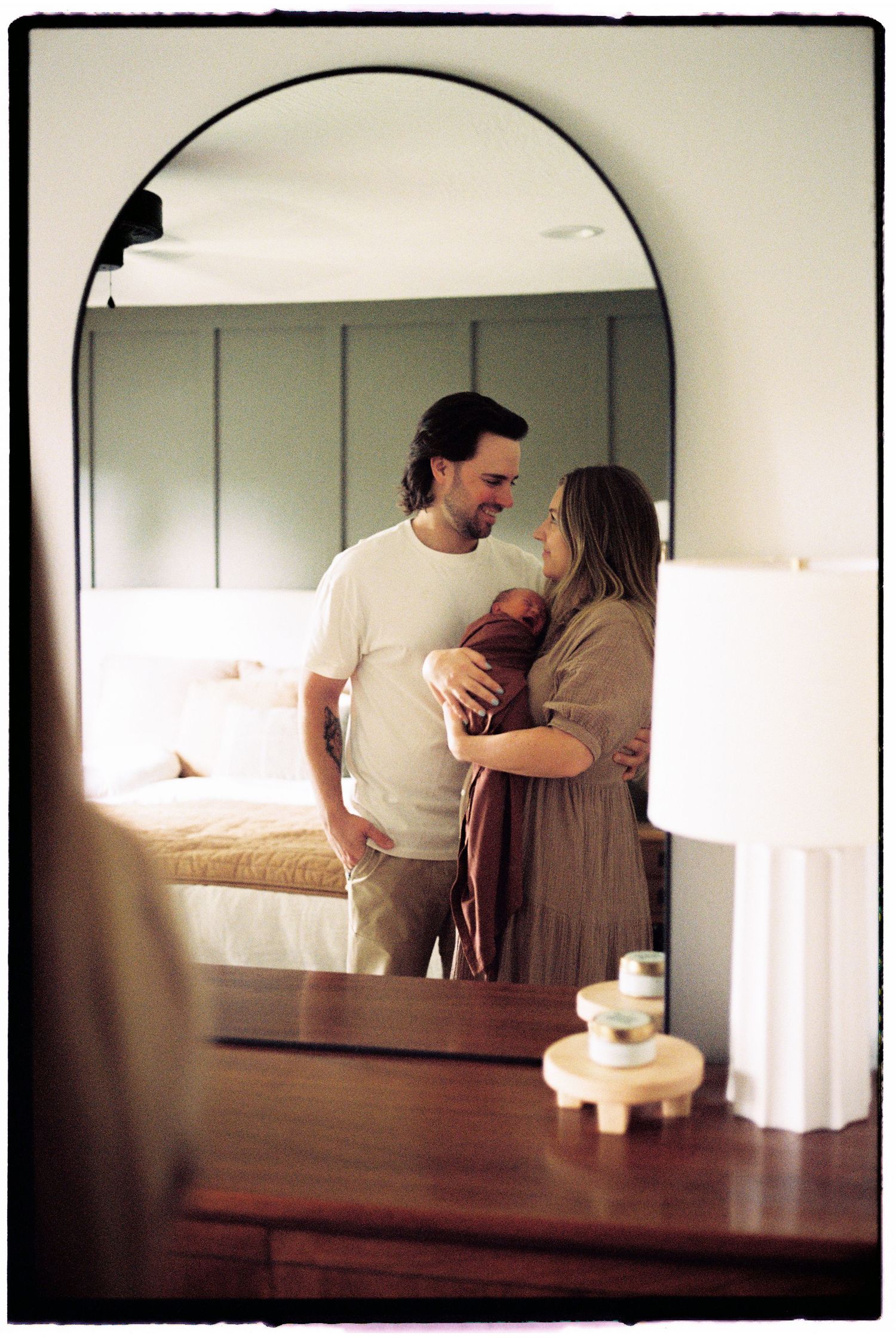 A tender moment captured in a bedroom mirror showing parents with their newborn baby.