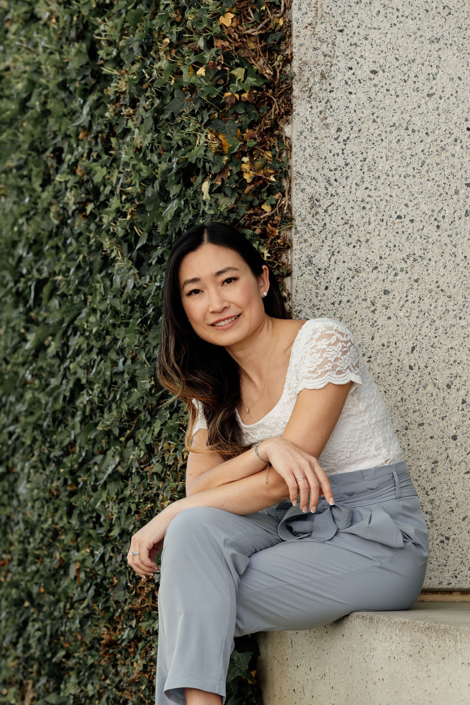 Woman in white lace top and gray pants sitting casually against wall with green ivy.