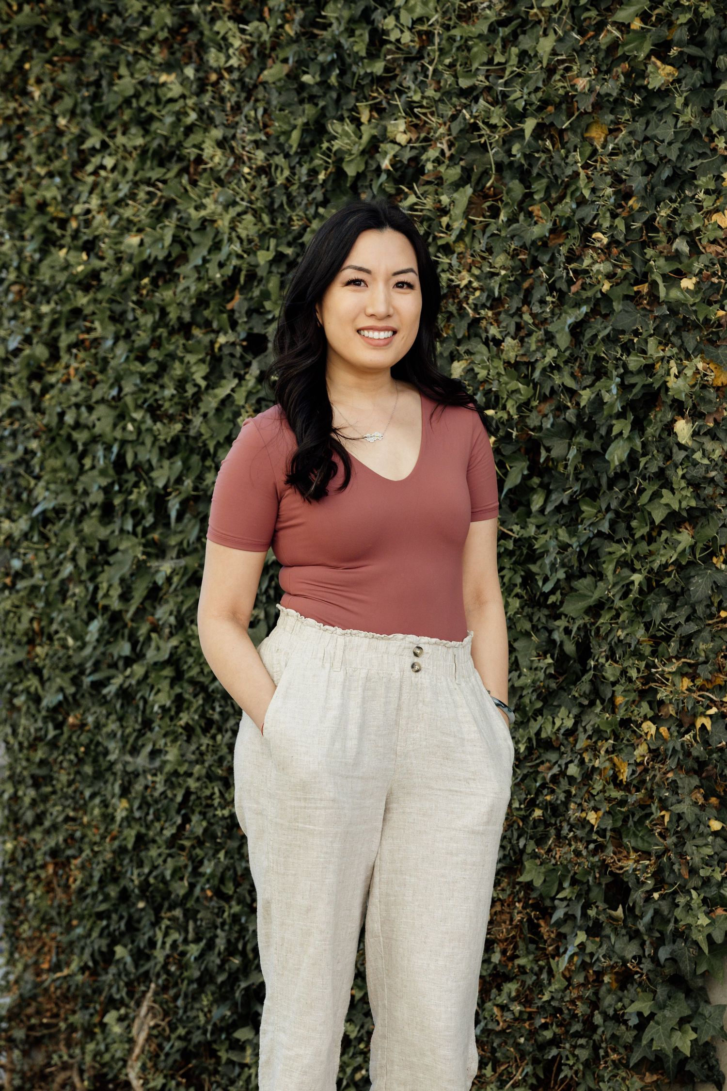 Woman wearing mauve top and beige pants posing against lush green vine wall.
