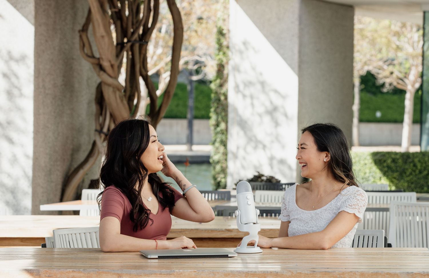 Two friends sharing laughter over coffee at an outdoor cafe table with a decorative tree in the background.
