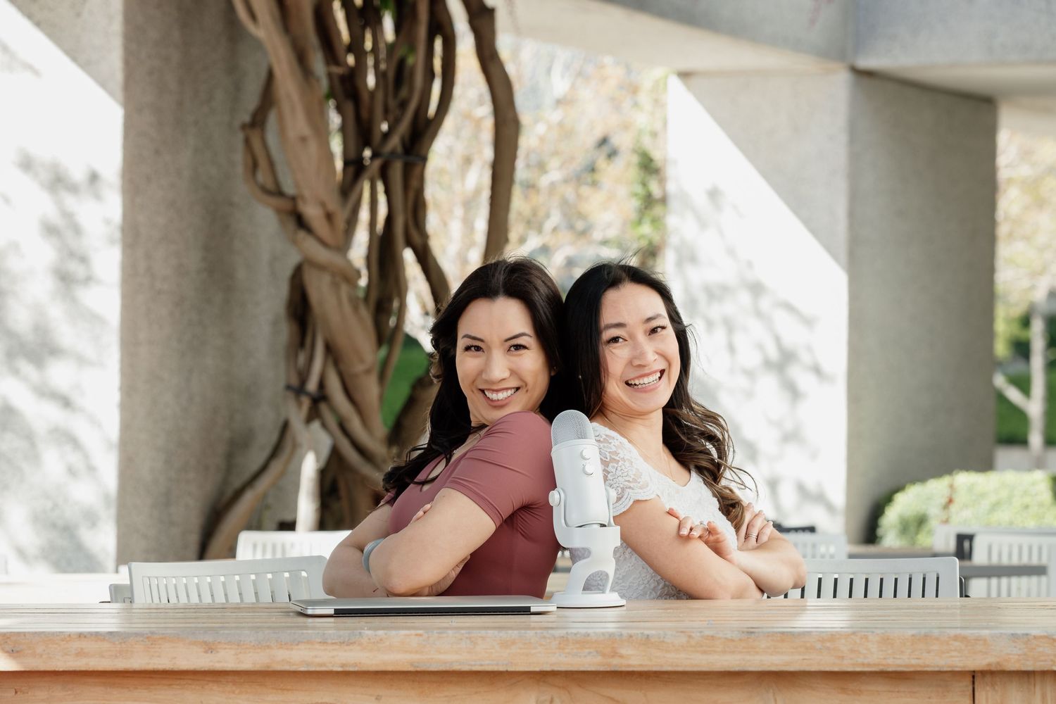 Two friends sitting back-to-back at an outdoor table while smiling warmly.
