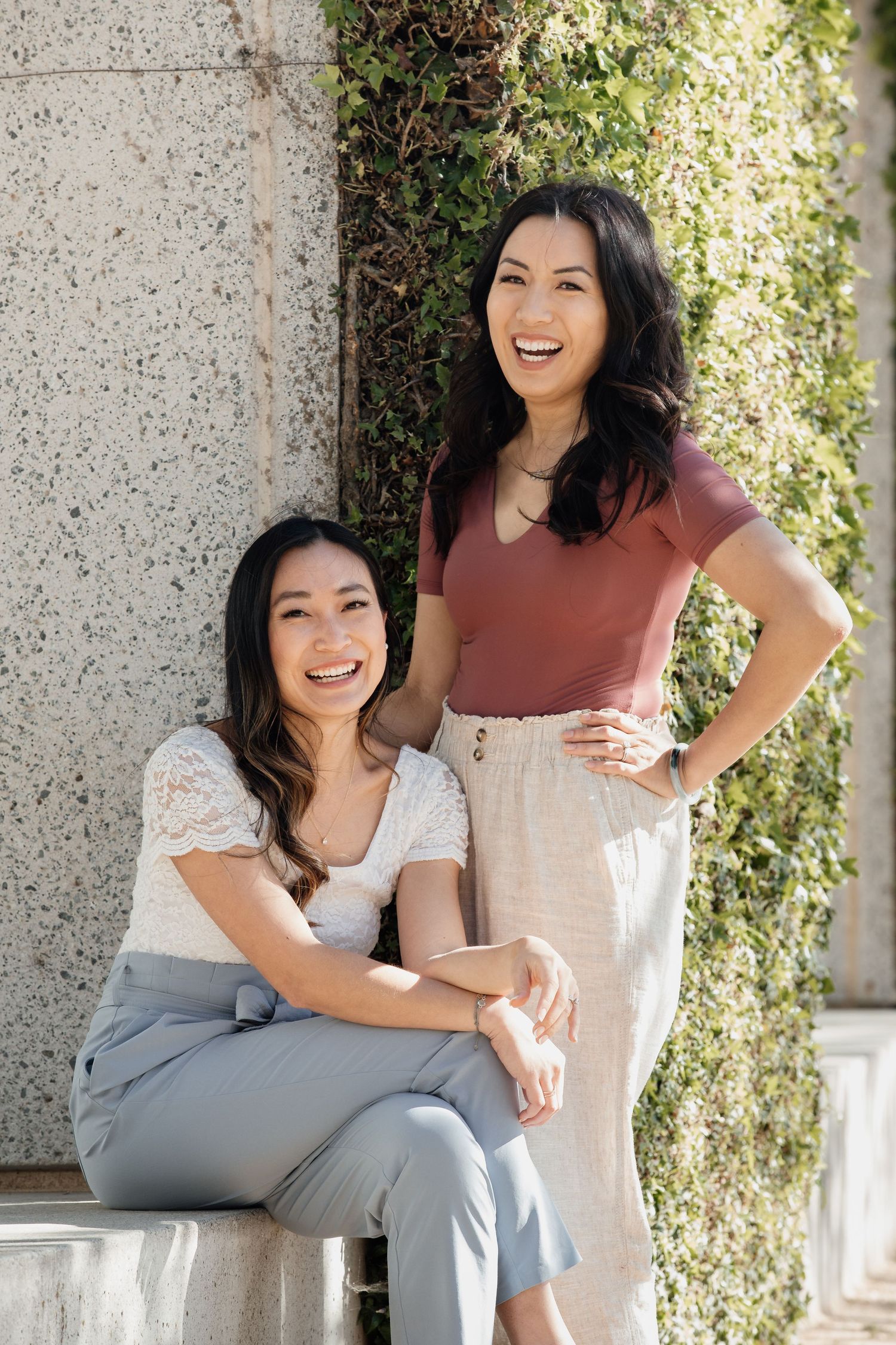 Two friends posing together by concrete wall with climbing vines in natural lighting.