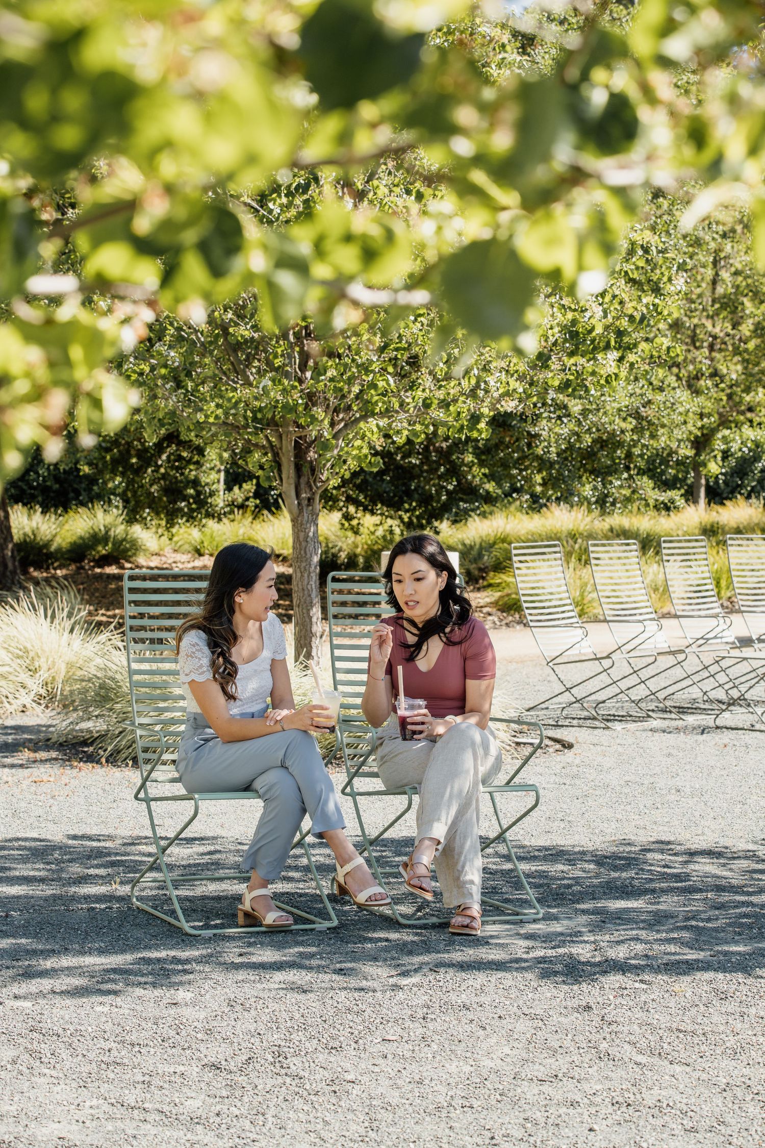 Friends enjoying conversation outdoors on metal chairs under dappled sunlight and trees.
