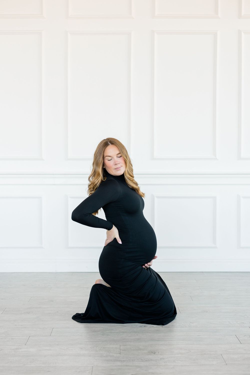 A pregnant woman in a black turtleneck dress poses gracefully on a light wooden floor in a bright white studio setting.