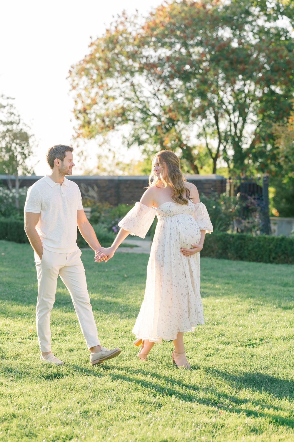 A couple in white outfits dance playfully together on a grassy hill during golden hour.
