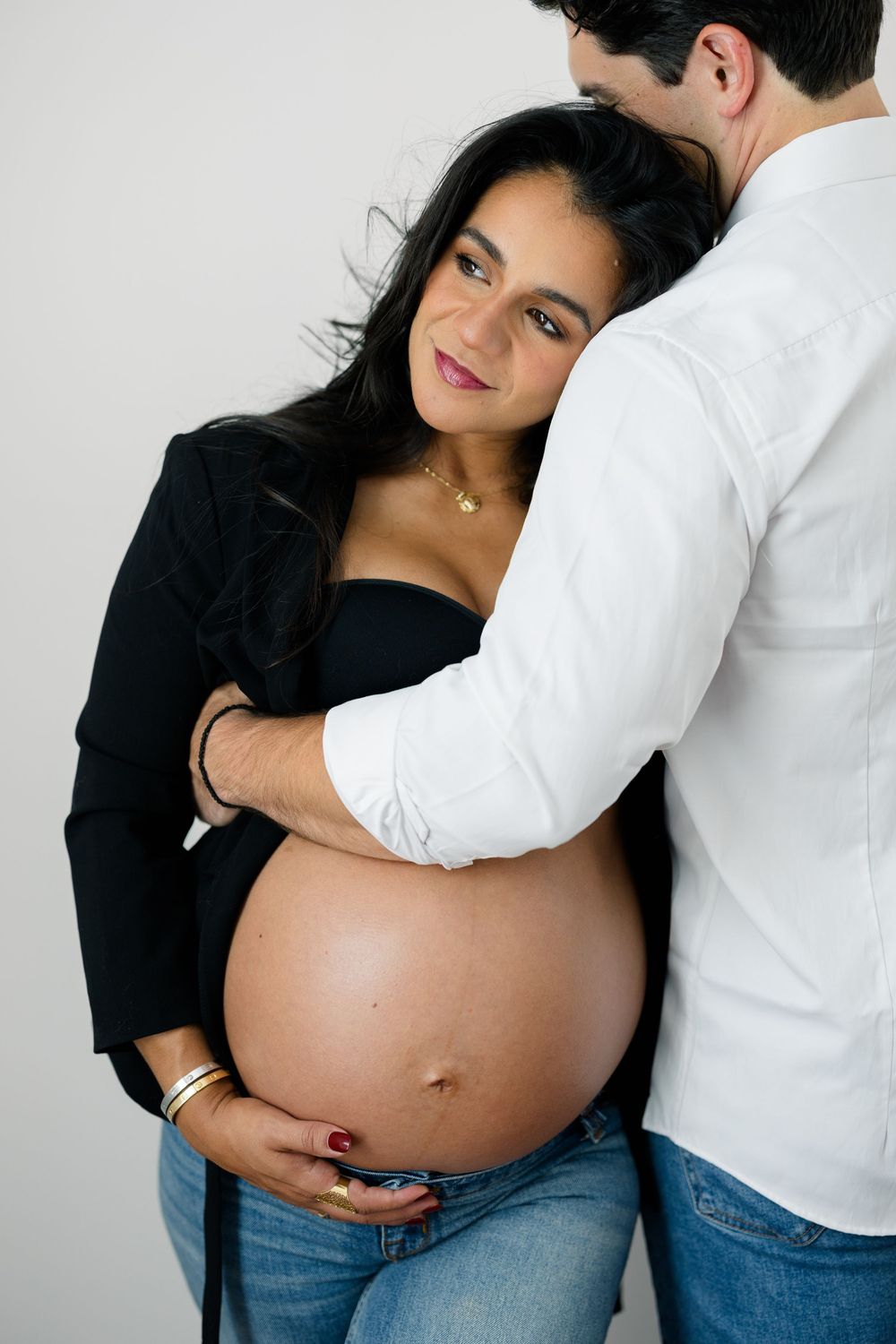 A couple shares an intimate moment as they embrace, with hands resting on a pregnant belly in a maternity photoshoot.
