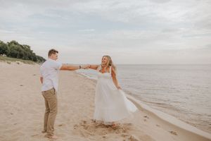 A romantic beach photoshoot with a couple dancing and twirling in white and tan outfits along the shoreline.