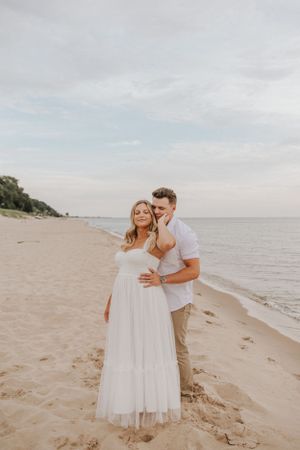 A couple shares playful moments on a sandy beach with waves and cloudy skies in the background.