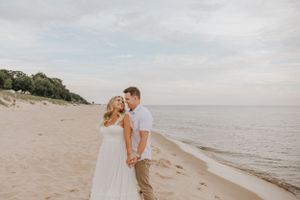 A couple embraces during a sunset beach photoshoot wearing coordinated white and tan outfits.