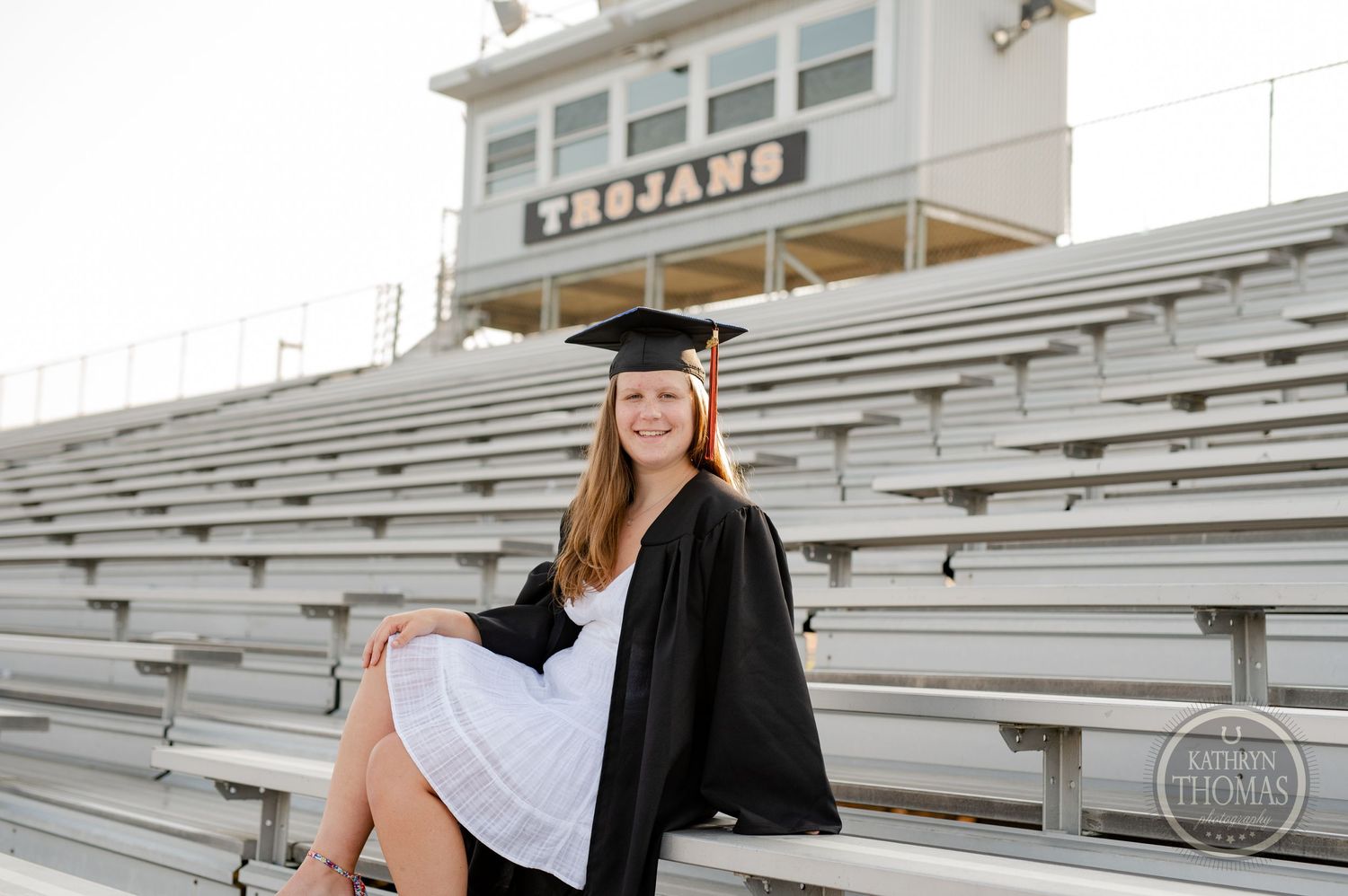 Graduate in cap and gown sits on stadium bleachers with white press box building in background.
