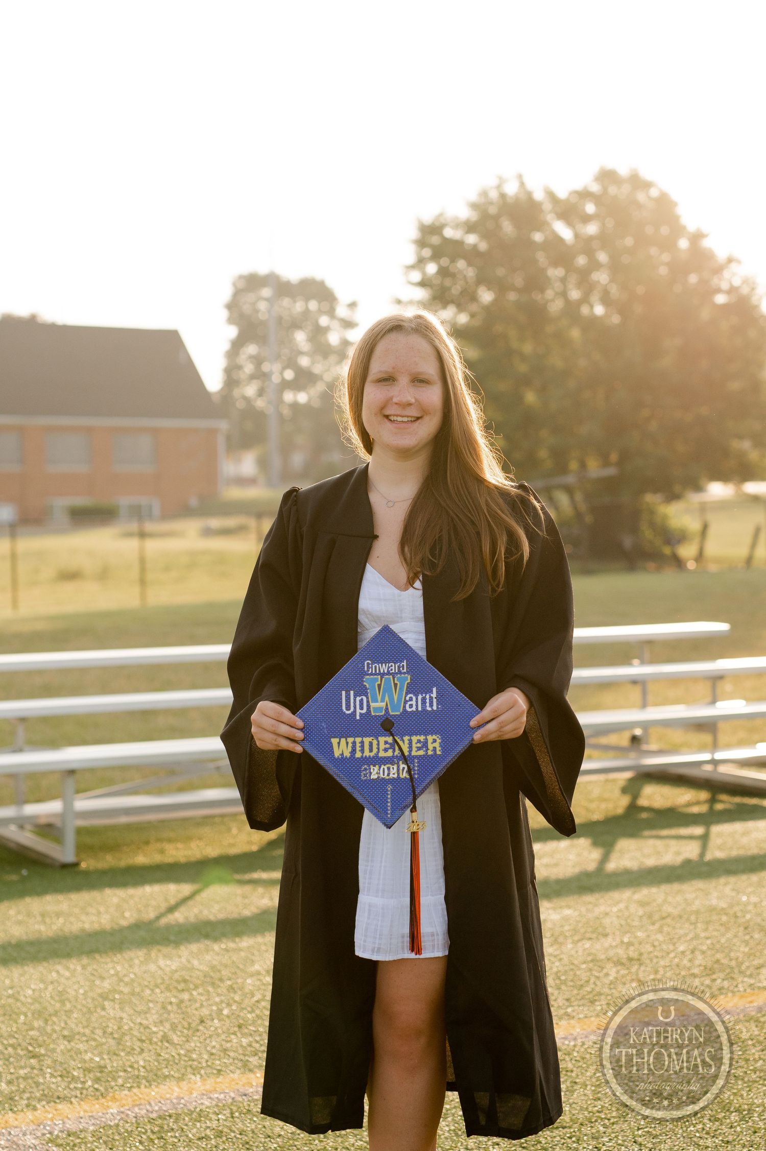 Graduate holds decorated cap while posing in graduation attire during sunset photo session.