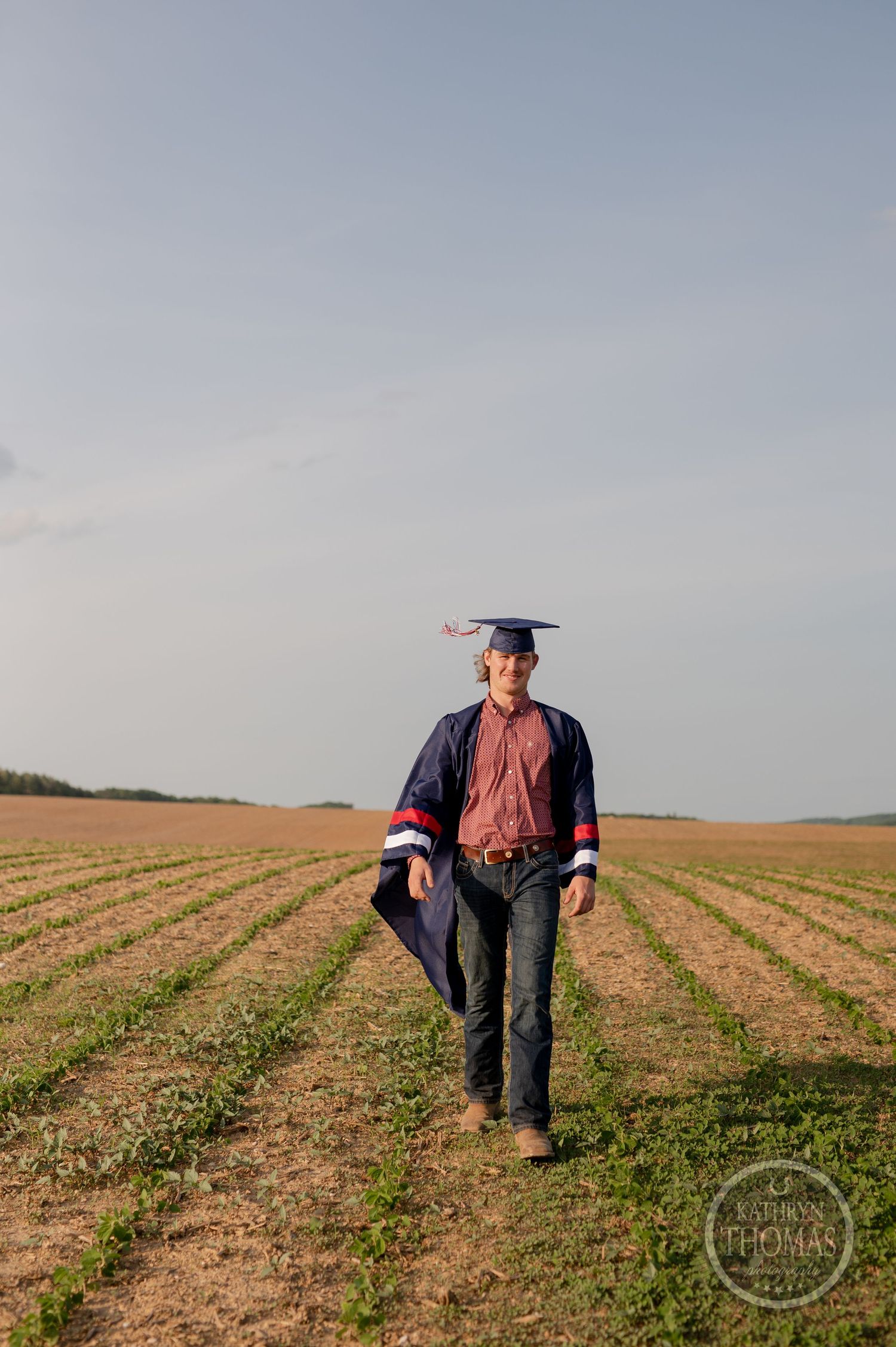 Graduate walks through planted field wearing cap and gown.