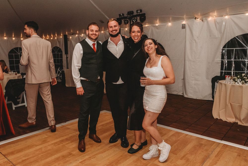 Wedding guests dance together on a wooden floor under white tent lighting during an evening reception celebration.