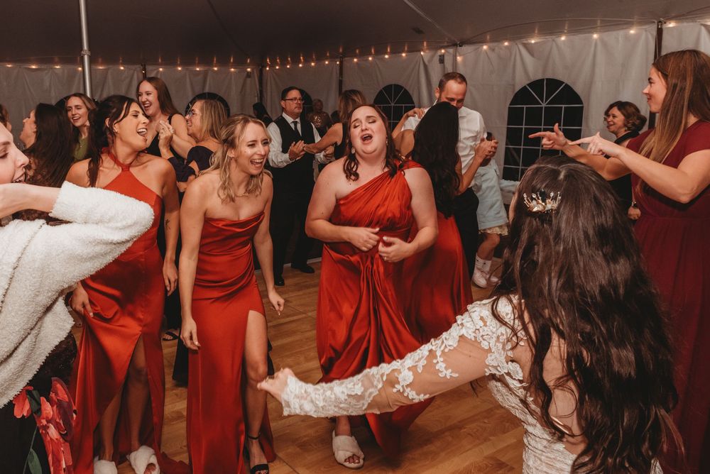 Wedding party guests in red dresses dancing and celebrating at a reception under string lights in a tent.