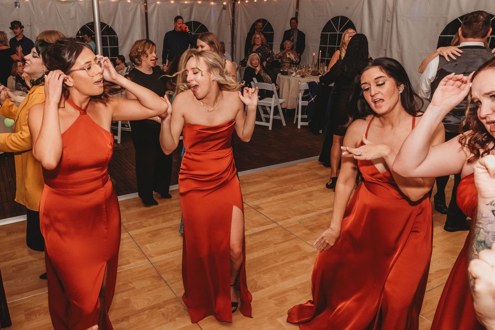 Bridesmaids in matching red dresses dance joyfully on a wooden dance floor at a wedding reception.