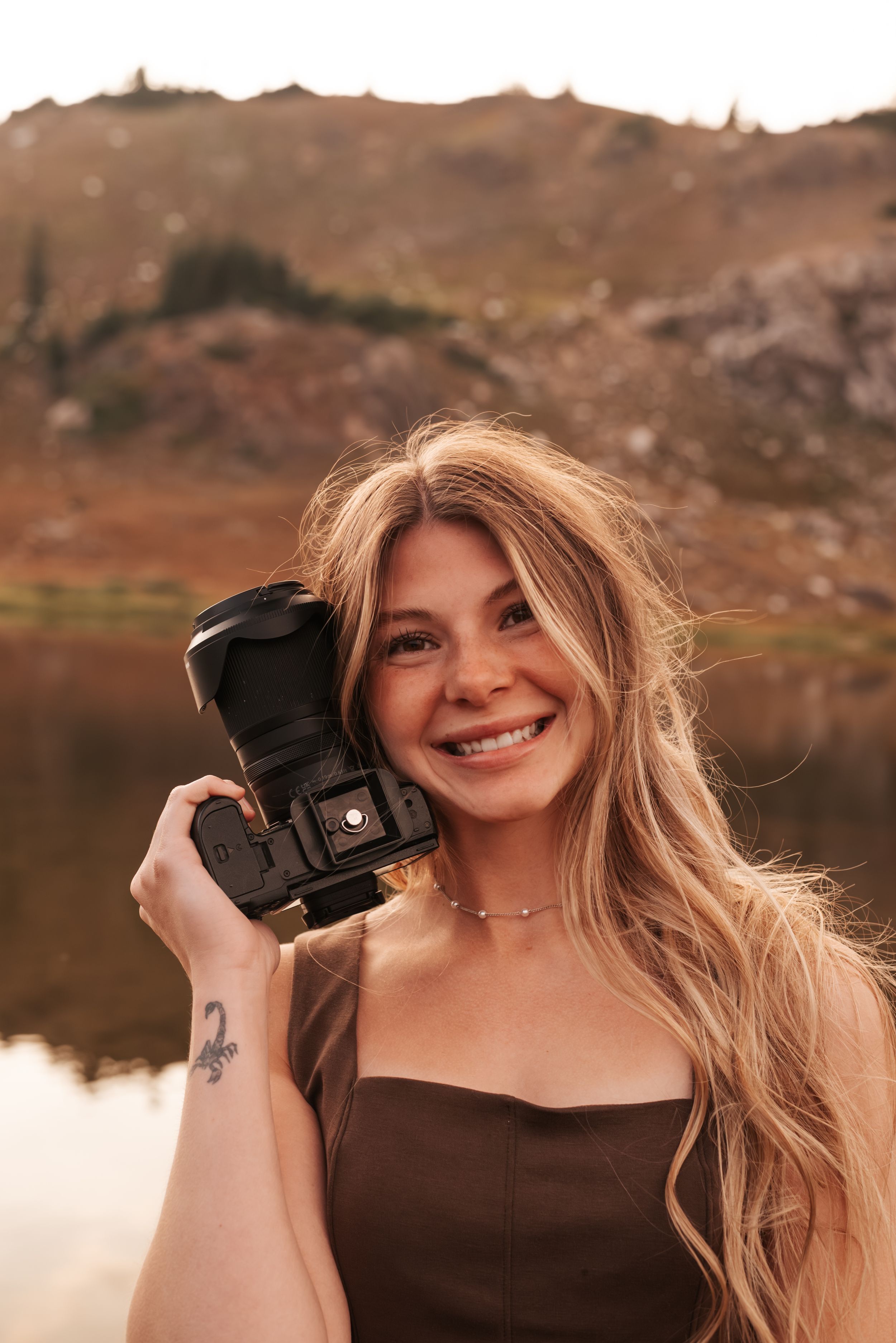 Woman holding camera smiles while wearing brown dress by lake.